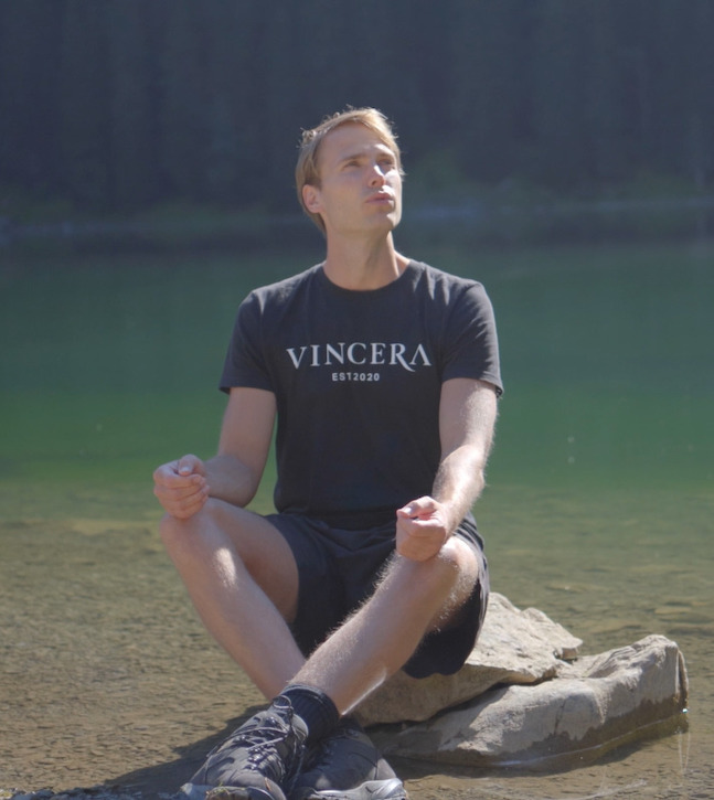 Een zittende man doet bij een meer een ademhalingsoefening in zijn Vincera-shirt