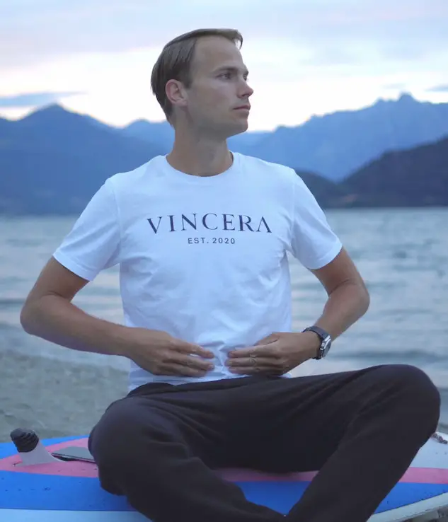 zittende man bij een meer legt twee handen op zijn buik voor buikademhaling, draagt wit vincera shirt